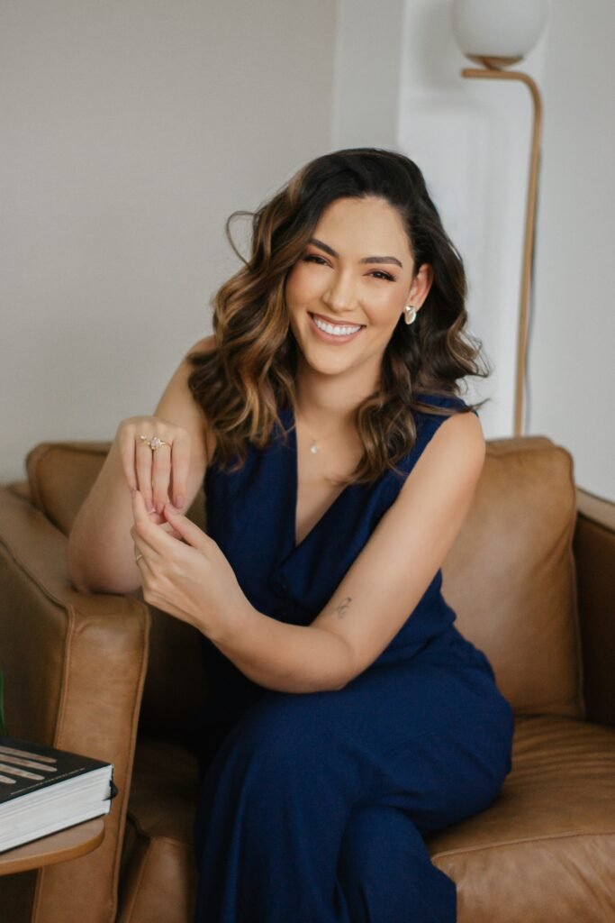 Terapeuta Júlia Rodrigues vestida de azul, sentada em um poltrona marrom de frente para a camera sorrindo