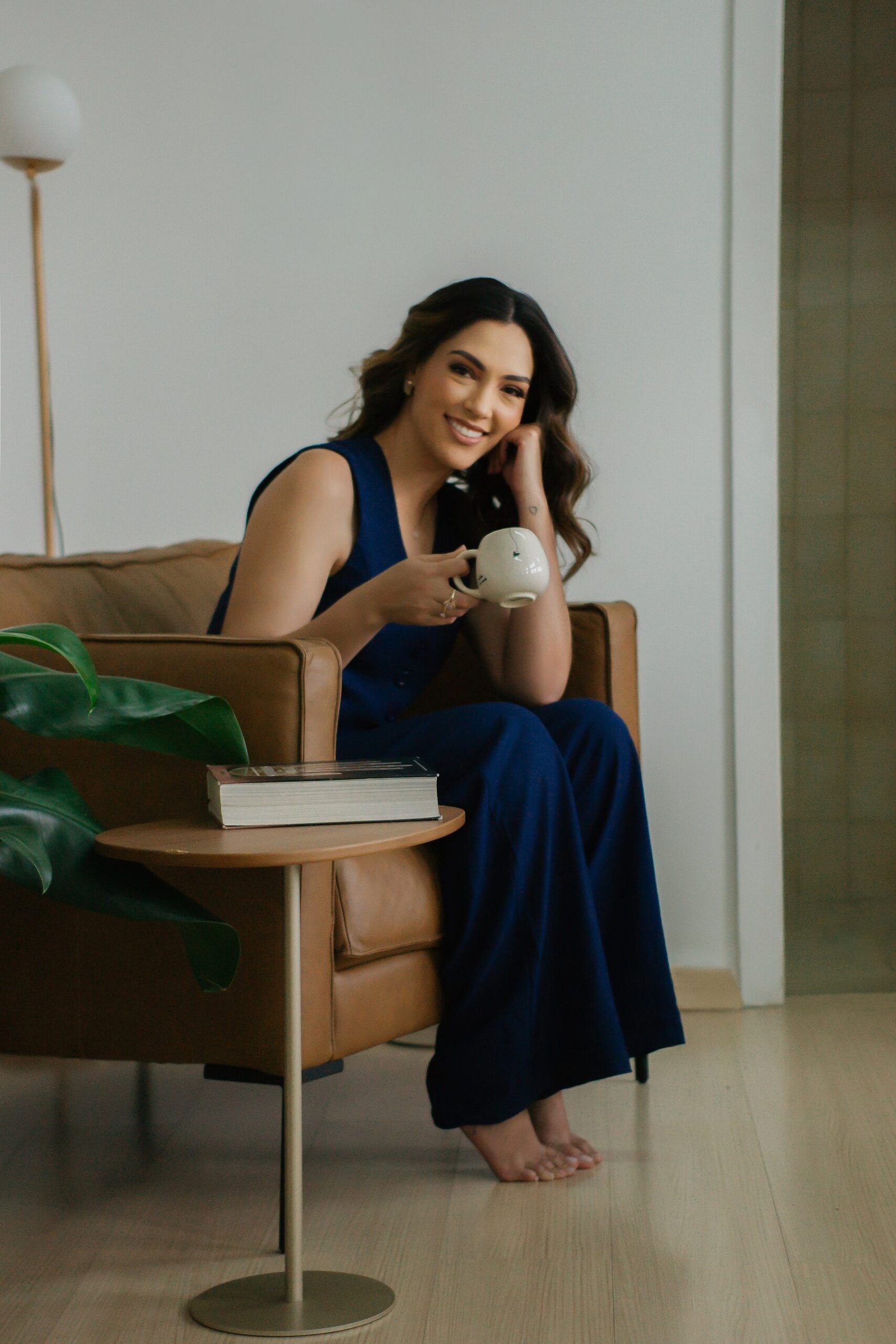 Júlia Rodrigues, mulher alta, vestida com um conjunto azul marinho, sentada em uma poltrona e com uma xicara de café na mão