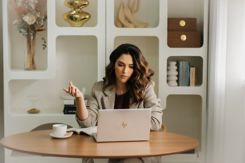 Terapeuta Júlia Rodrigues, que esta de cabelos soltos, blazer cinza, blusa marrom no fundo, senta e com um notebook a frente, realizando uma sessão online.